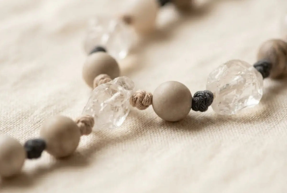 Hand-knotted natural-stone talisman strand on cream linen: clear quartz and neutral stone beads spaced along a hand-dyed cotton cord, a small hand-tied knot between every bead, no metal clasp, wire, or glue.