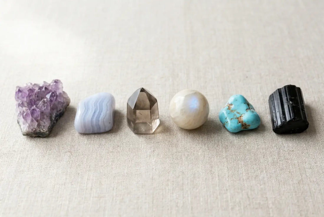 Six calming crystals for anxiety on linen: raw amethyst, blue lace agate, a smoky quartz point, moonstone, turquoise with brown matrix, and black tourmaline.