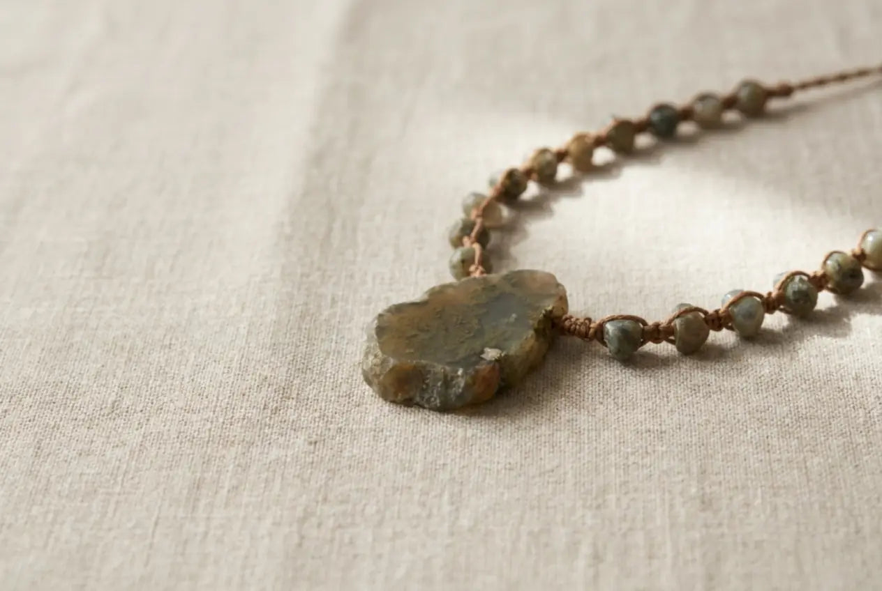 A single edition-of-one hand-knotted natural-stone talisman necklace with a raw-edged focal stone on knotted cord, resting on oatmeal linen â the Ã  la luck quiet-luxury aesthetic.