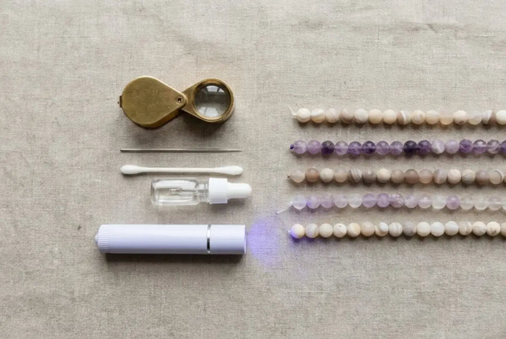 Crystal-jewelry authentication kit on linen — a brass 10x loupe, steel needle, cotton swab, glass dropper bottle, and a violet-glowing UV lamp beside natural stone bead strands — the home-verification toolkit from à la luck