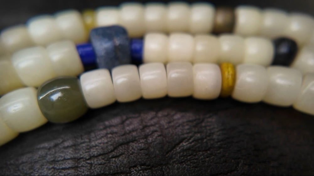 Macro of The Spark #29, a hand-knotted 108-bead mala with cream disc-cut camel bone and cultural marker beads — lapis lazuli, a grey-green stone, and an ochre disc — resting on dark leather, a la luck edition-of-one