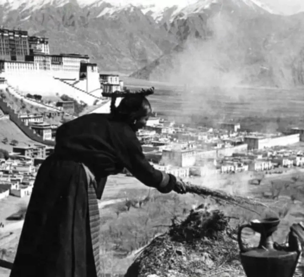 Vintage black-and-white photograph of a Tibetan noblewoman in traditional chuba and headdress performing a juniper-incense sangsöl ritual on a high stone wall, with smoke rising over the Potala Palace and the Lhasa valley