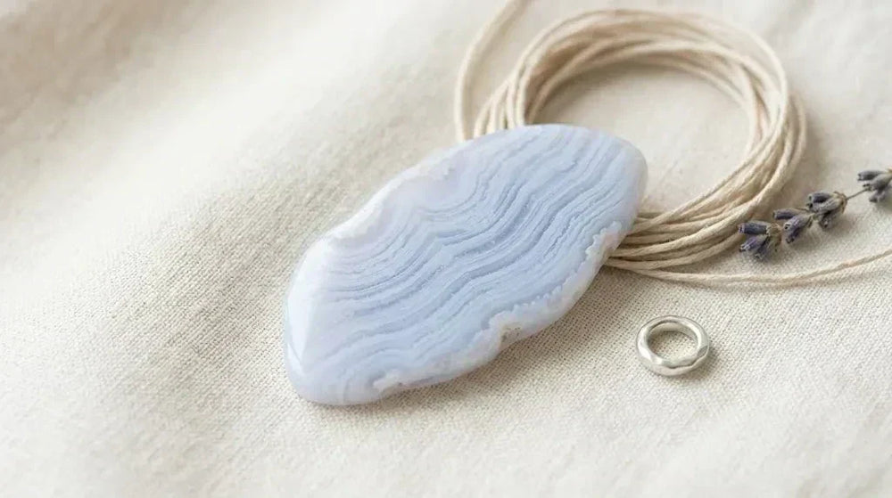 Pale blue lace agate tablet showing soft horizontal lace banding on cream linen, with a coil of natural cream cotton cord, a sprig of dried lavender and a small silver jump ring — featured image for the blue lace agate bracelet and throat chakra guide