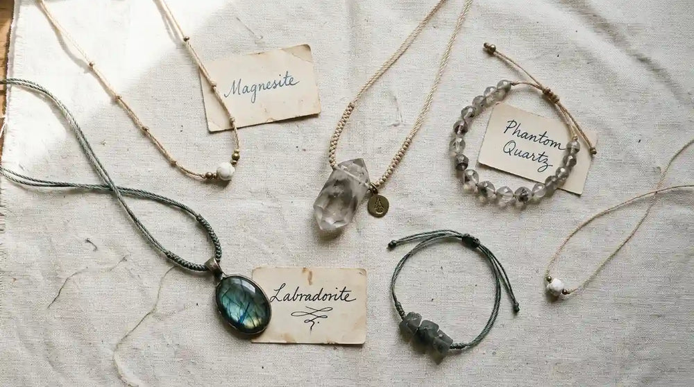 Flat-lay of five handmade crystal talismans on cream linen — hand-knotted cord work in macro detail. Three paper cards label stones in plain mineralogical names: Magnesite, Phantom Quartz, Labradorite. Soft morning light, considered composition.