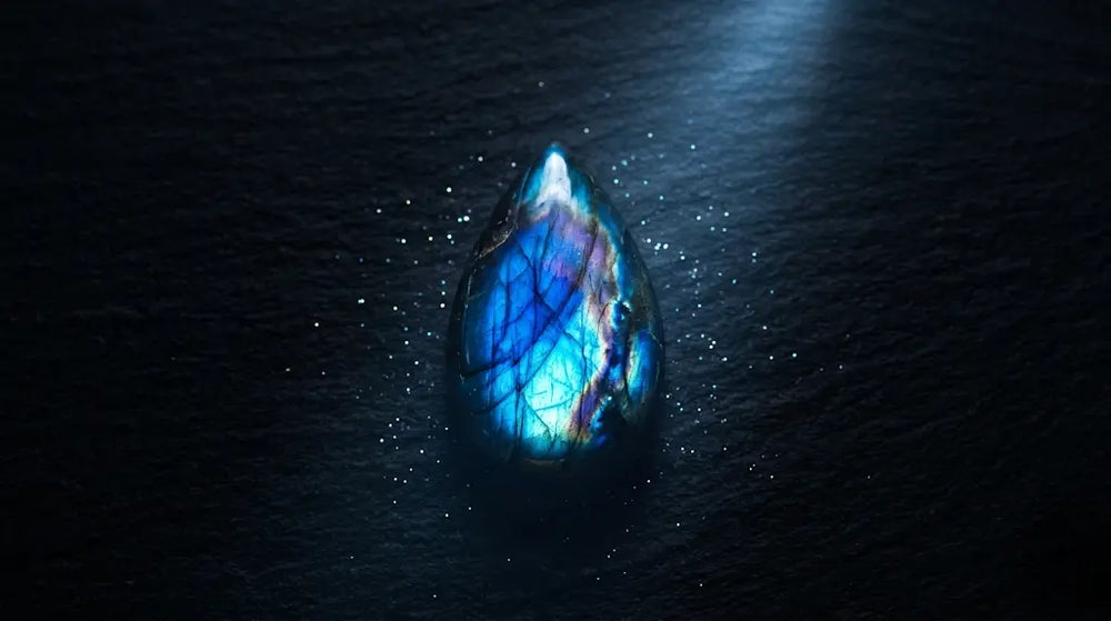 Macro of a teardrop labradorite cabochon flashing electric blue and rainbow labradorescence on dark slate — featured image for the labradorite meaning and stone-of-intuition guide