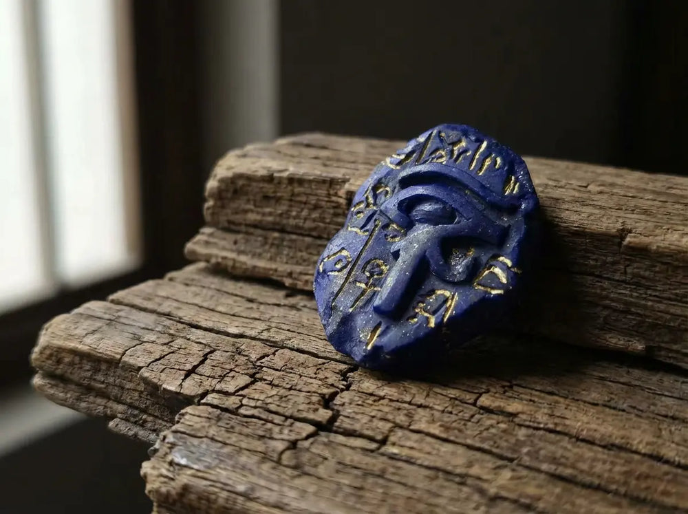 Carved lapis lazuli amulet shaped as the Eye of Horus / Wedjat with gold-inlaid hieroglyph cartouche resting on weathered split timber by a window — featured image for the lapis lazuli Eye of Horus meaning article