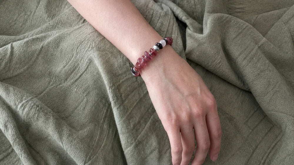 #34 worn close on the wrist over olive-green linen, showing softly irregular Strawberry Quartz beads, Garnet, Moonstone, Sunstone, metal spacers, and a fixed fit