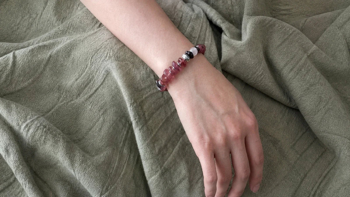#34 worn close on the wrist over olive-green linen, showing softly irregular Strawberry Quartz beads, Garnet, Moonstone, Sunstone, metal spacers, and a fixed fit