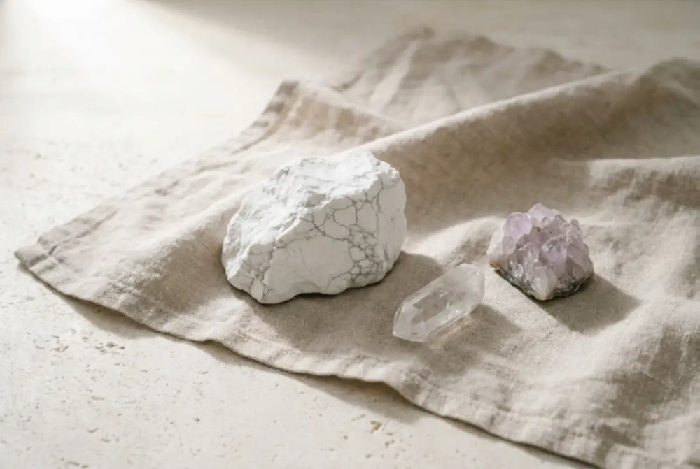 Three natural clarity crystals on oatmeal linen â a white magnesite chunk with grey veining, a clear quartz point, and a pale amethyst cluster â placed with calm negative space, illustrating a guide to crystals for mental clarity and focus.