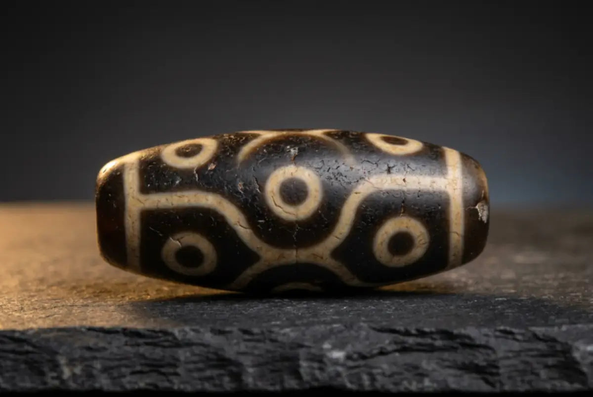 Antique Tibetan dzi bead — an etched banded-agate barrel bead with a dark brown-black ground and pale concentric 'eye' circles, showing age weathering and patina, on dark slate. The honest material behind the 'heaven's bead.'