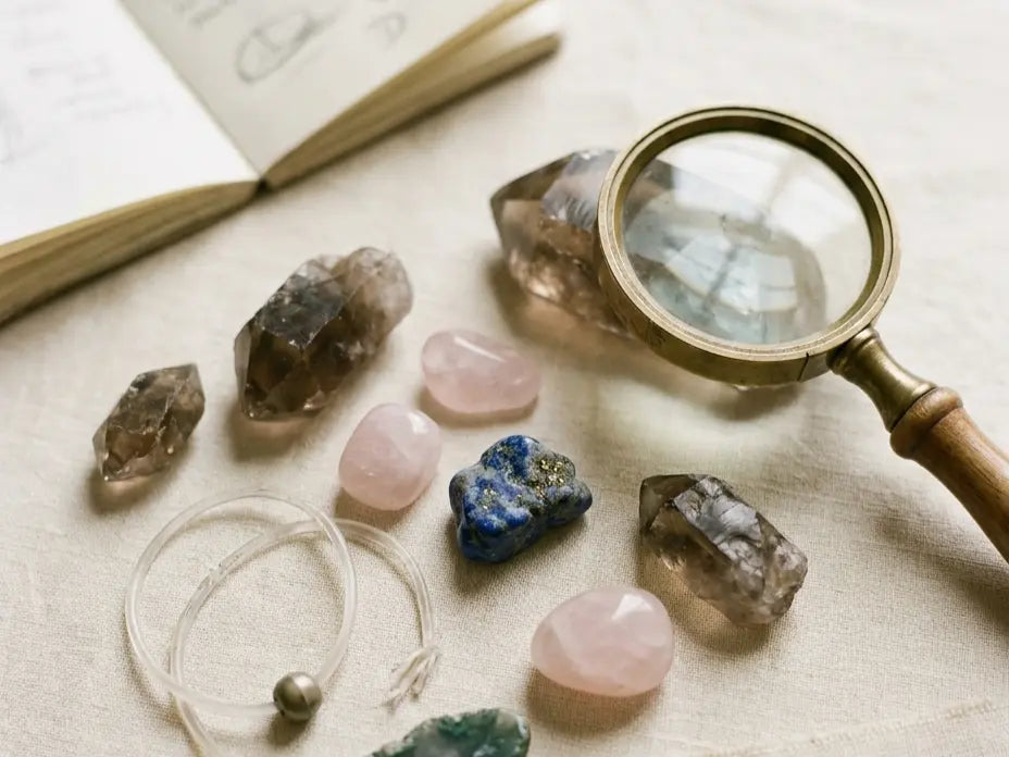Editorial flatlay of scattered raw crystals, a brass magnifying glass, an open handwritten notebook, and a coiled frayed elastic cord with one loose bead on warm cream linen — the visual shorthand for reading crystal brand reviews carefully.