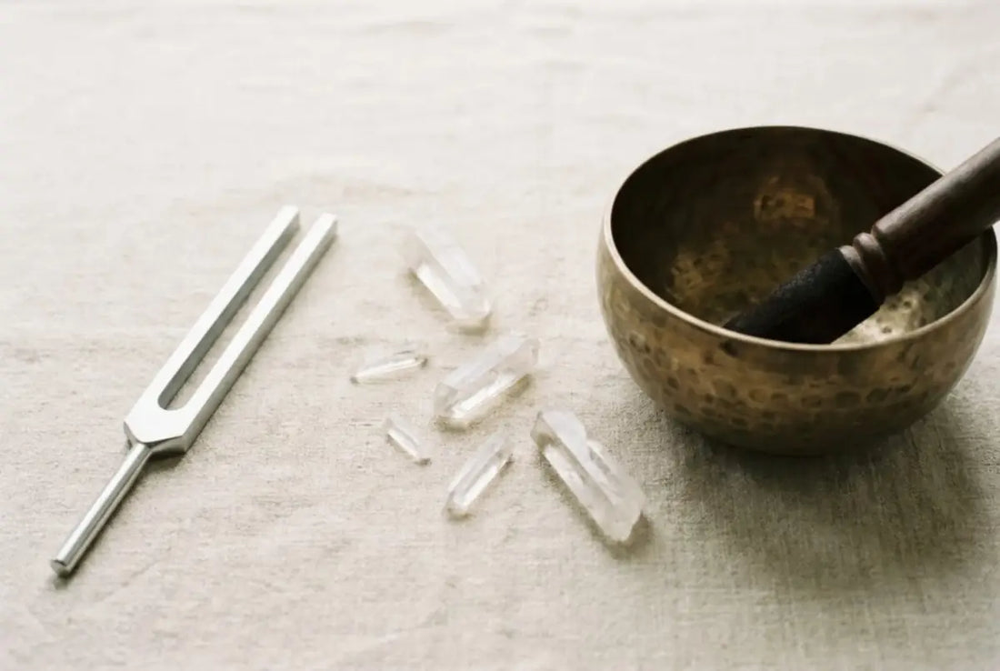 A steel tuning fork and a bronze singing bowl side by side on oatmeal linen with clear quartz points, comparing two crystal sound-cleansing tools