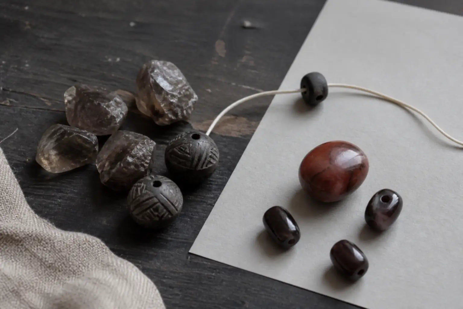 Raw smoky-brown crystal pieces, carved dark clay beads, smooth red and brown beads, and ivory cord arranged on a worn maker's table