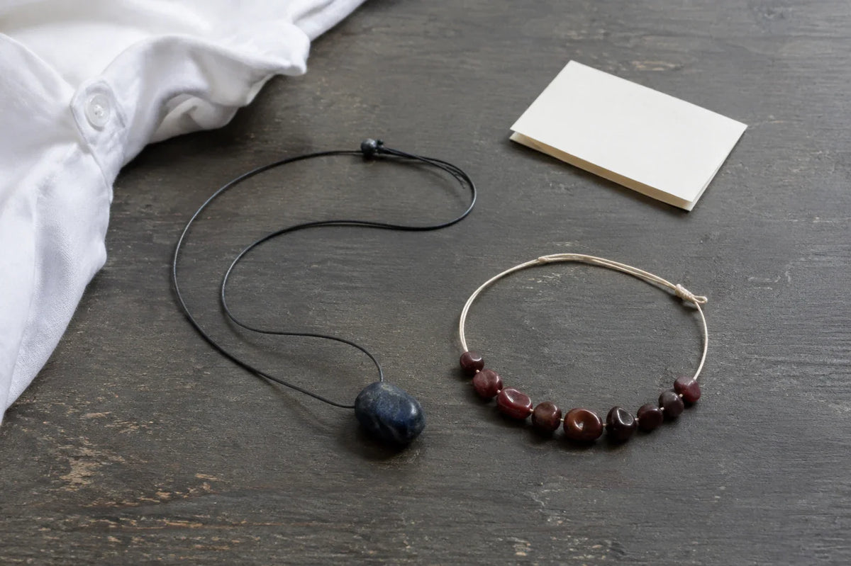 AI-generated still life of a blue stone pendant, red-bead bracelet, white cotton shirt and blank folded note on worn gray wood