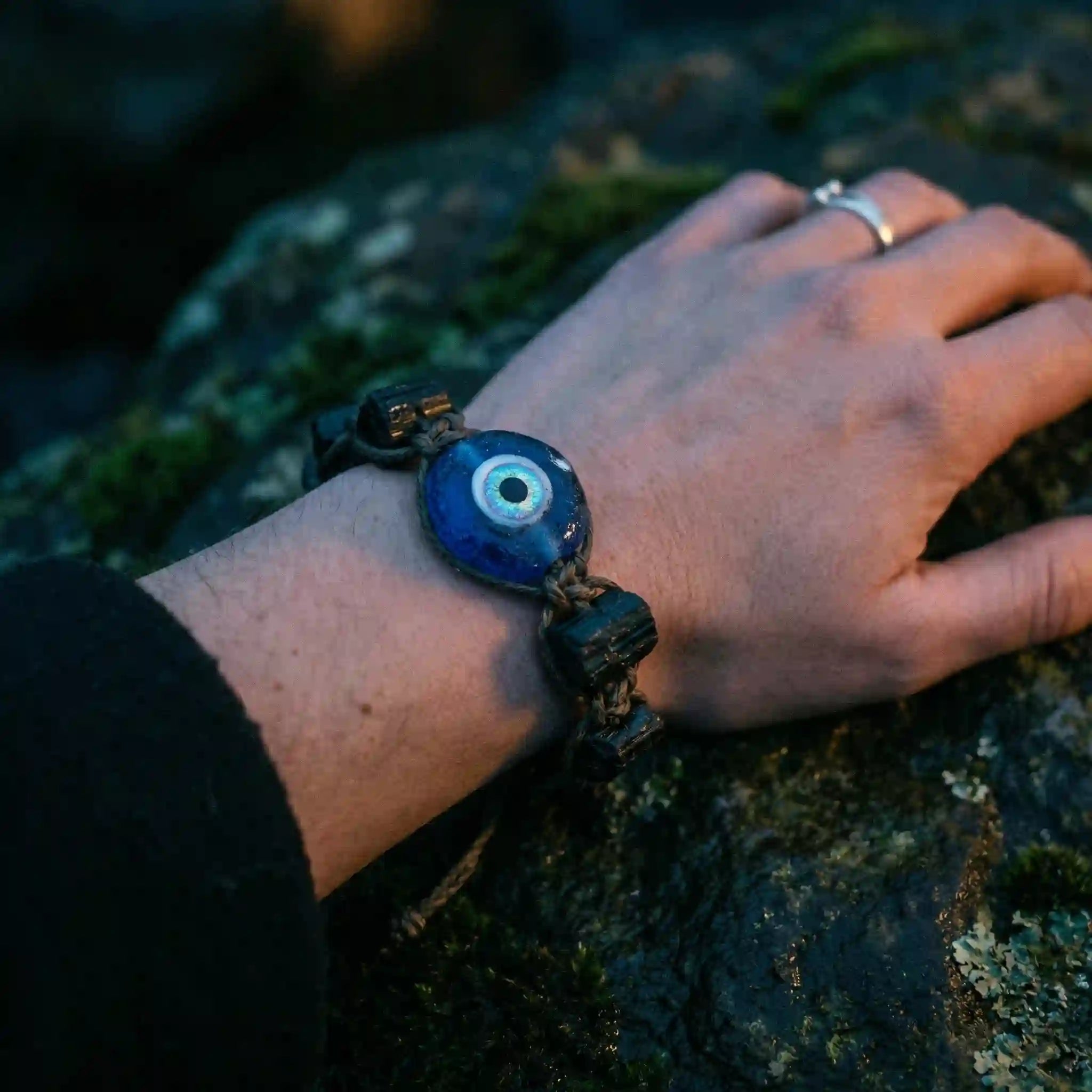 a hand wearing protection almulet with Evil Eye bead and Black Tourmaline stones resting on a dark ancient rock covered in moss