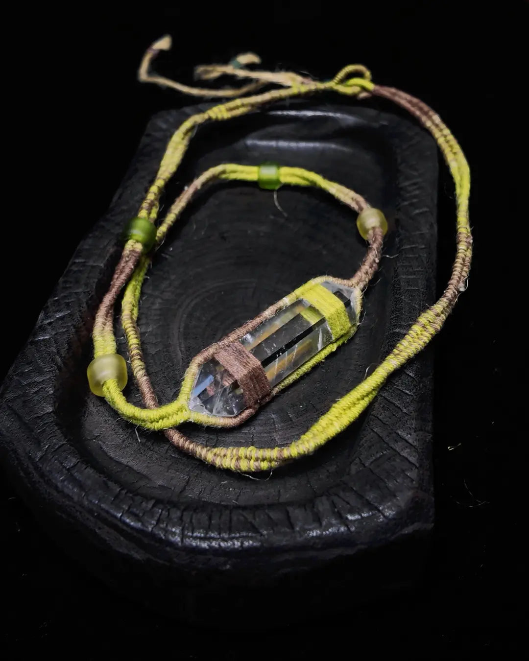 The Mindfulness #01 Himalayan clear quartz wrap shown flat on charred wood — full edition-of-one piece showing the raw quartz centerpiece, green silk and plant-dyed cotton thread, and small glass beads at the wrap loop.