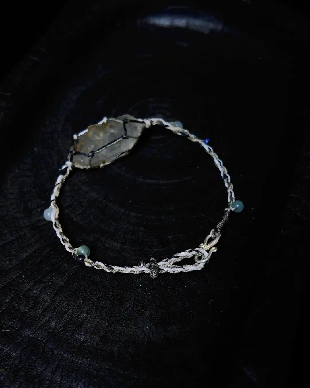 The Alchemist #09 Himalayan smoky quartz macrame bracelet by à la luck — full edition-of-one piece flat on dark charred wood showing the raw smoky quartz focal in macrame waxed thread cage with blue jadeite and white jade accent beads.