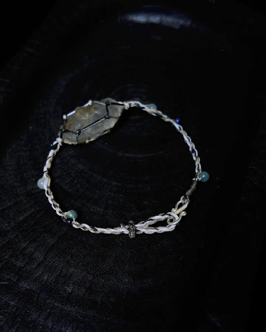The Alchemist #09 Himalayan smoky quartz macrame bracelet by à la luck — full edition-of-one piece flat on dark charred wood showing the raw smoky quartz focal in macrame waxed thread cage with blue jadeite and white jade accent beads.