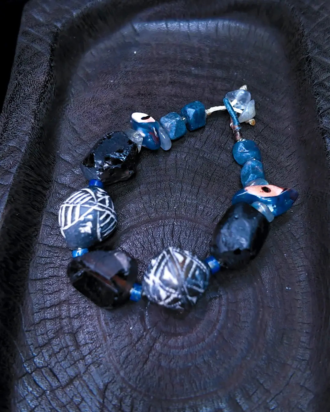 The Ancestor #31 smoky quartz and Mali clay grounding bracelet, full flat lay on dark wood showing complete bead arrangement with blue stone chips and evil eye bead