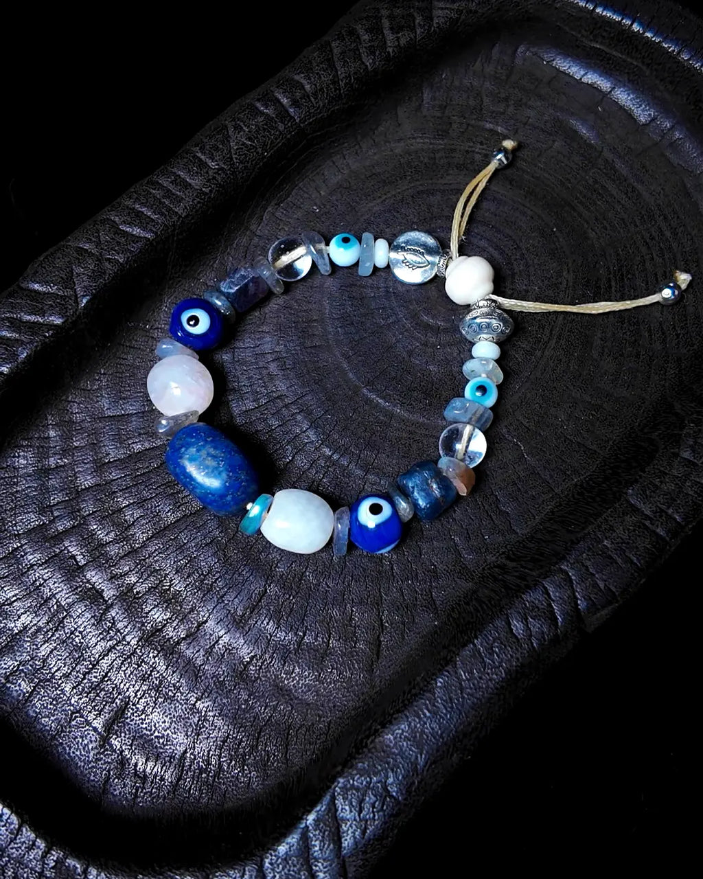 The Augur #33 lapis lazuli and blue moonstone vision bracelet, full flat lay on dark wood showing complete blue-gemstone arrangement with evil eye glass
