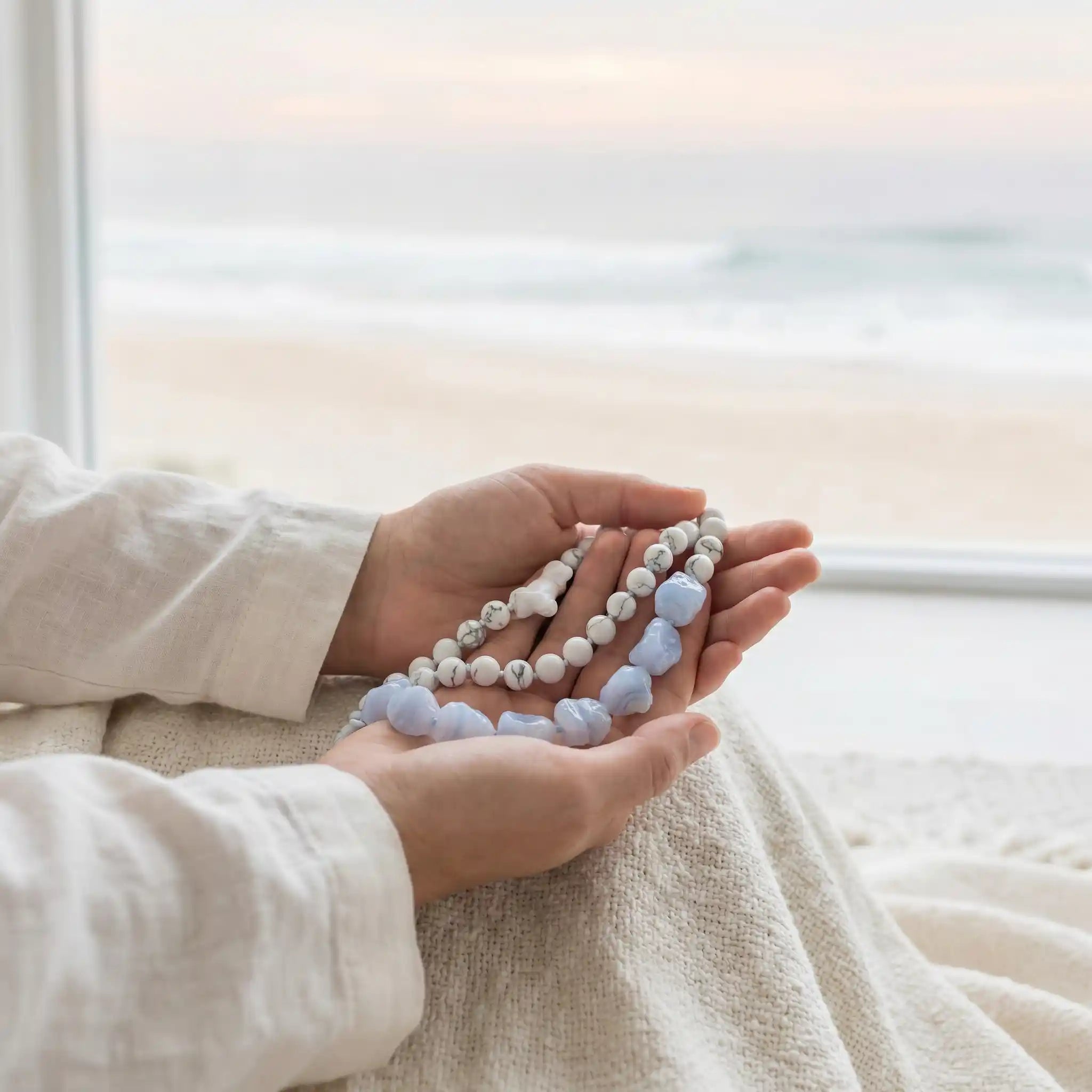 Person holding a string of white howlite and blue lace agate rawstone beads in front of a window with soft light.