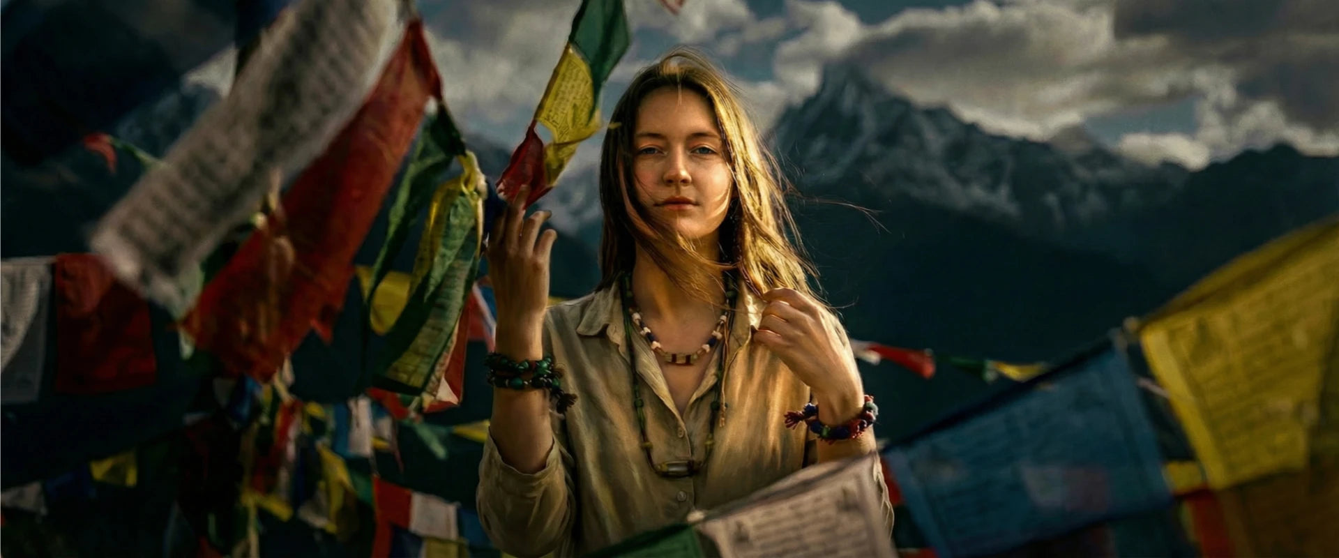 Woman standing amidst colorful prayer flags with mountains in the background wearing colorful talismans